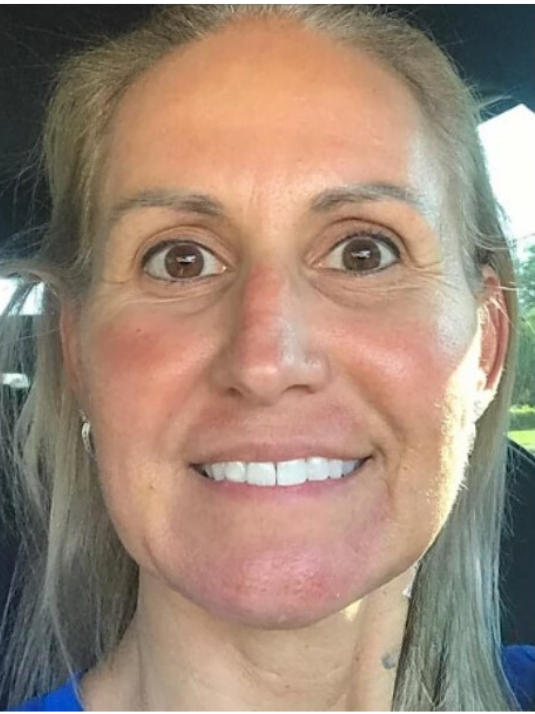 A smiling person with long blonde hair and blue shirt, looking directly at the camera in a brightly lit setting.