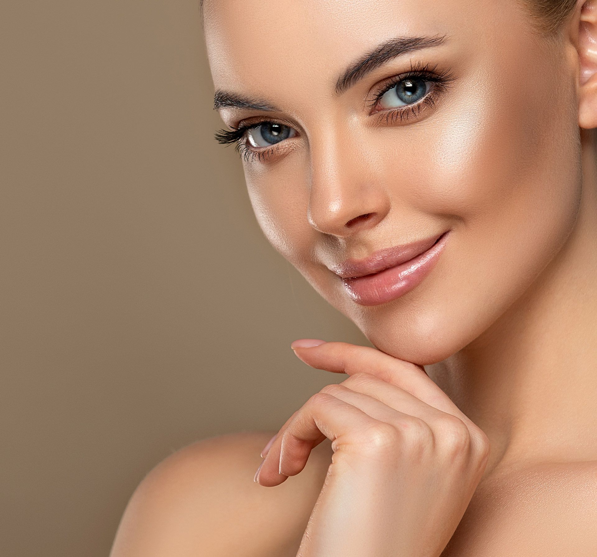 Woman with glowing skin smiles, hand on chin, against a neutral background.