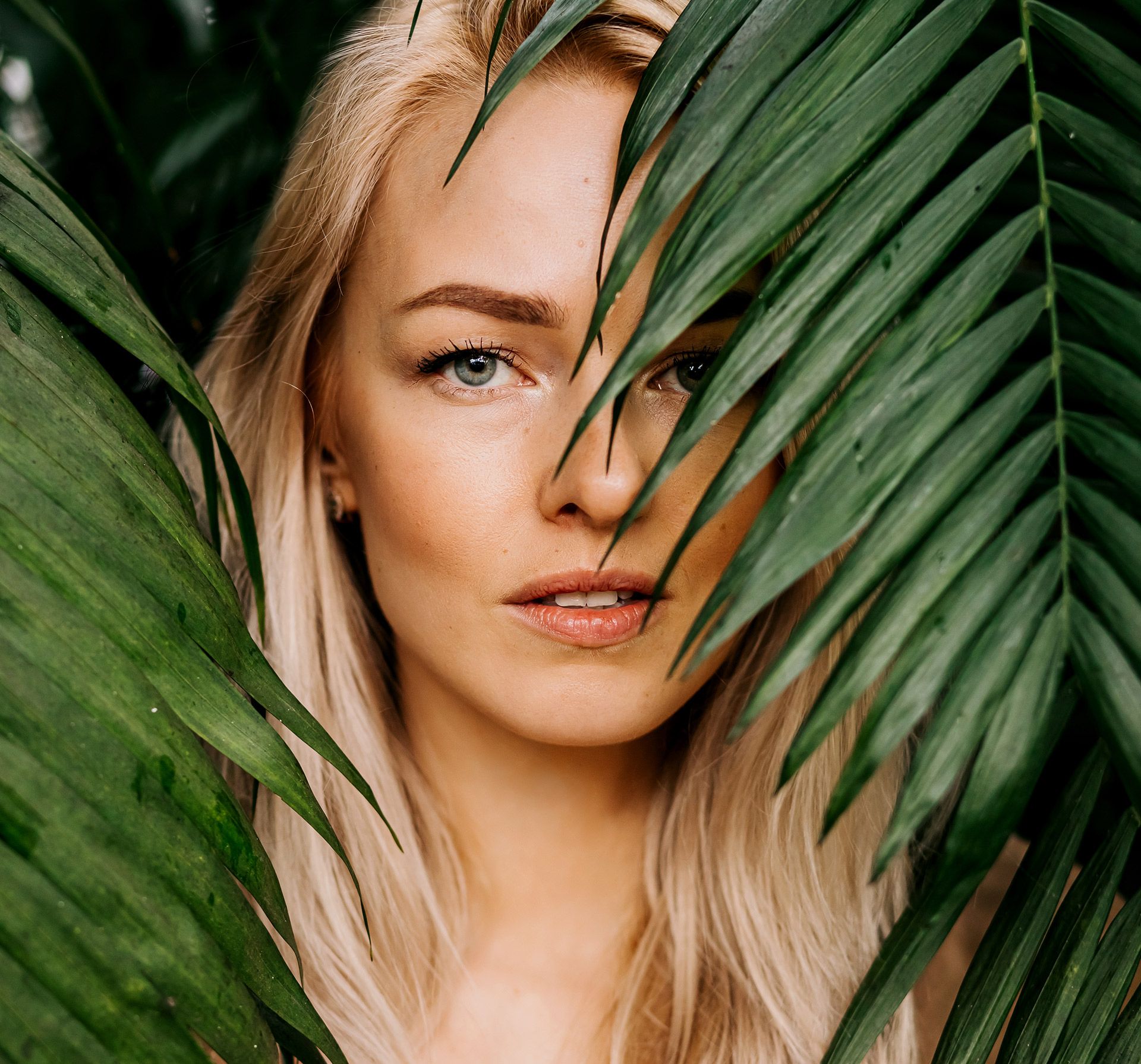 Blonde woman peeks through large green leaves, looking directly at the viewer with a neutral expression.