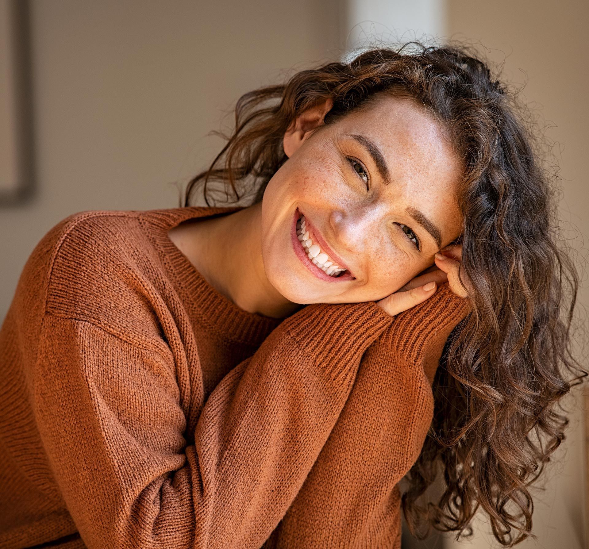 Woman with curly brown hair smiles, leaning on arms, wearing a rust-colored sweater.