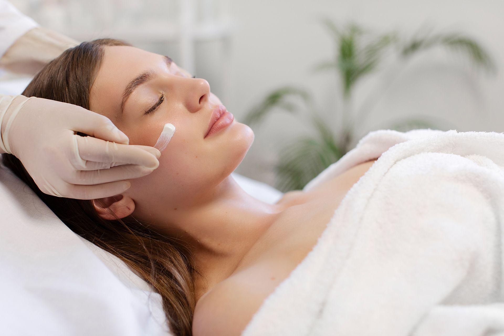 A gloved hand applies cream to a person's face while they lie down during a spa treatment.