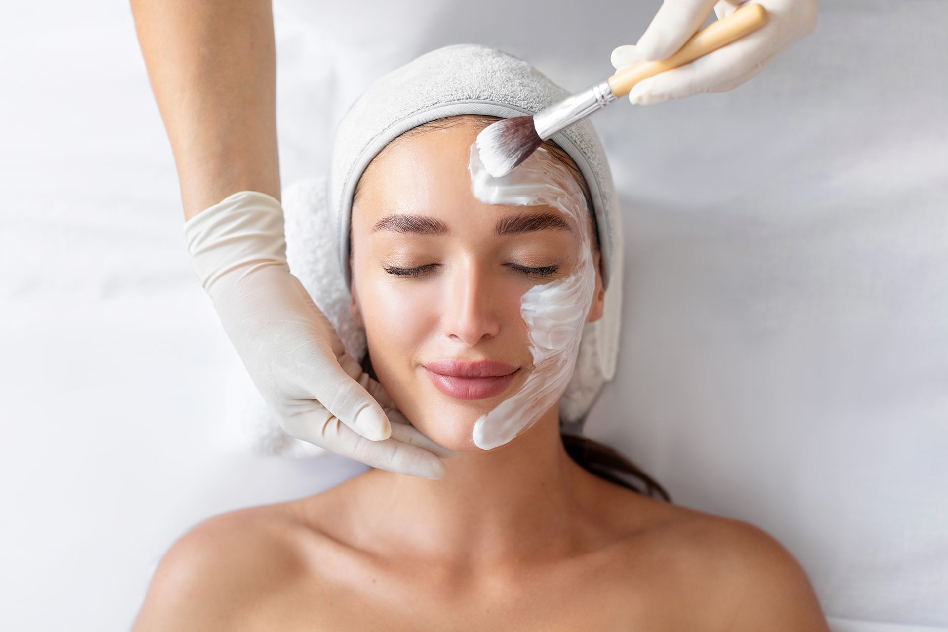 Person receiving facial treatment; cream mask being applied with a brush by gloved hands, in a spa setting.