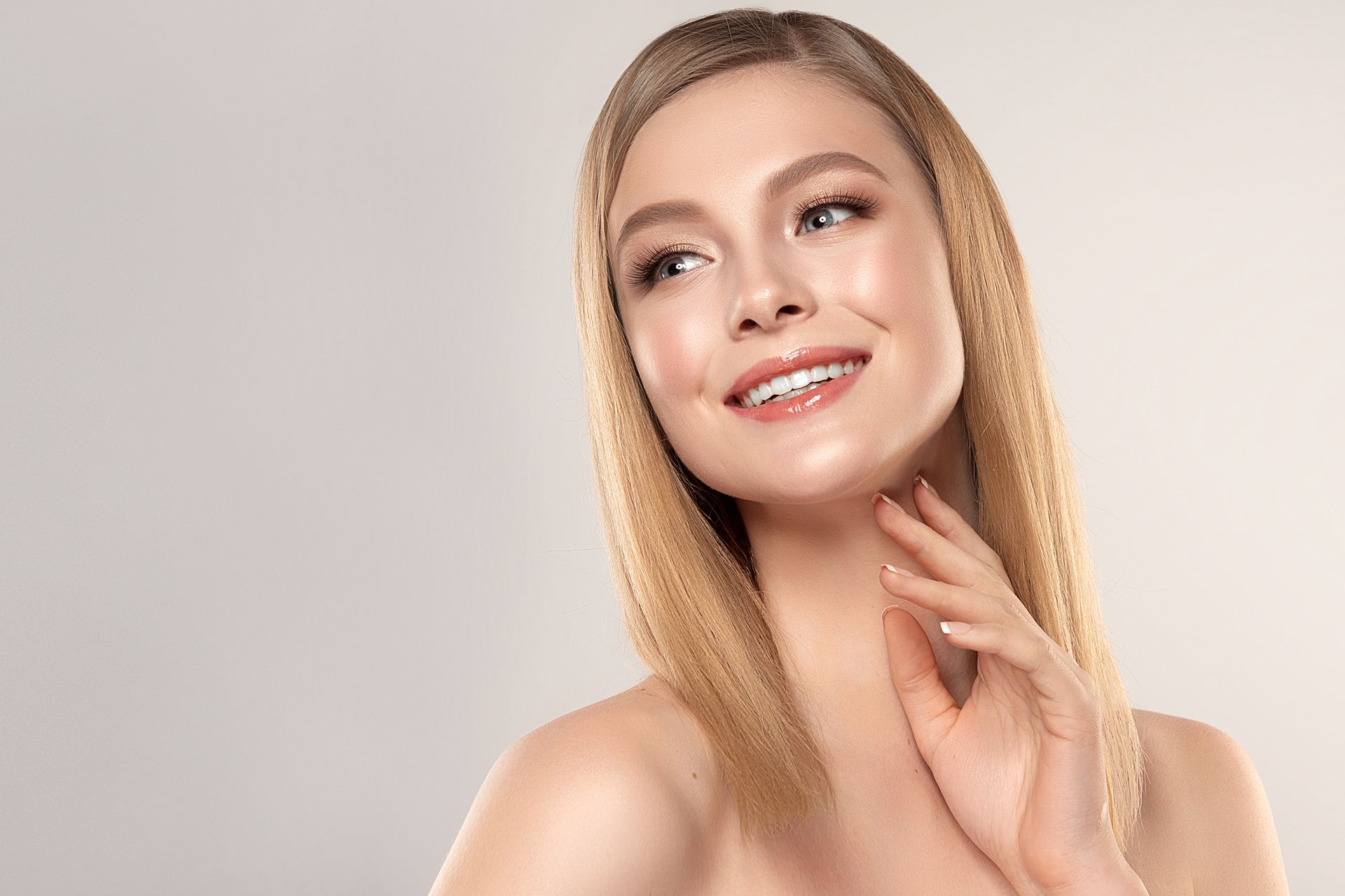 Woman with straight blonde hair smiles, touching neck. Beige background.