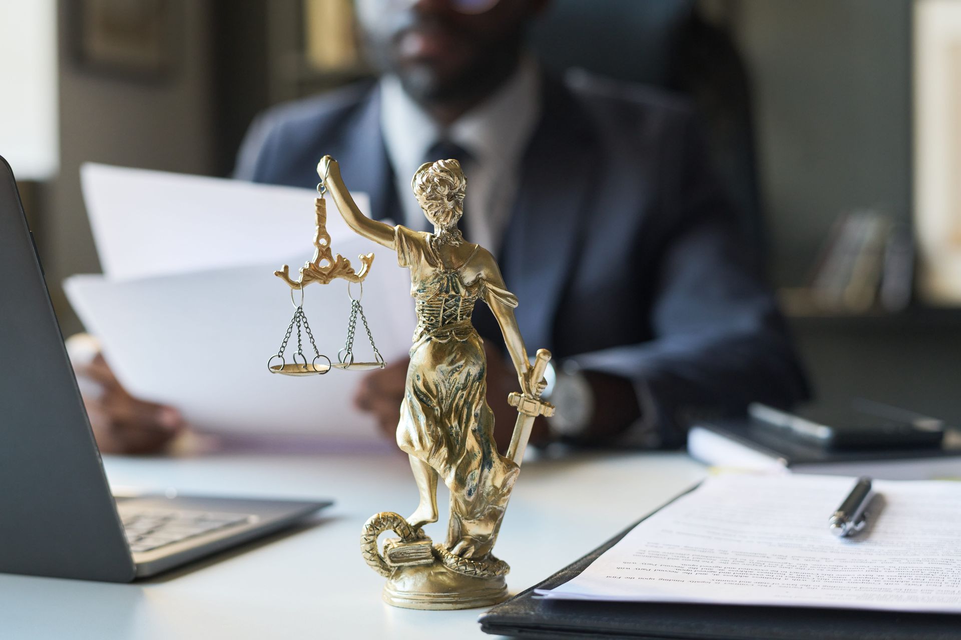A small gold scales of justice statue sits on a desk in front of an out-of-focus person in a suit holding legal documents.