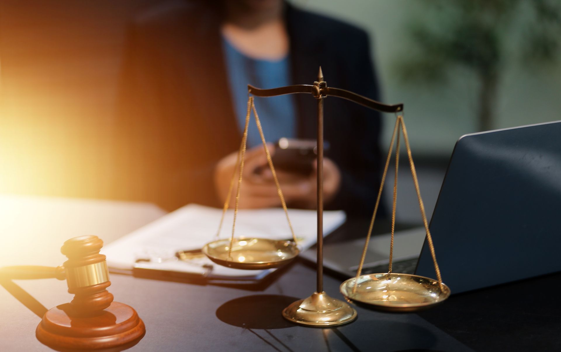 A judge's gavel and a set of brass scales of justice on a desk with a blurred figure in the background.