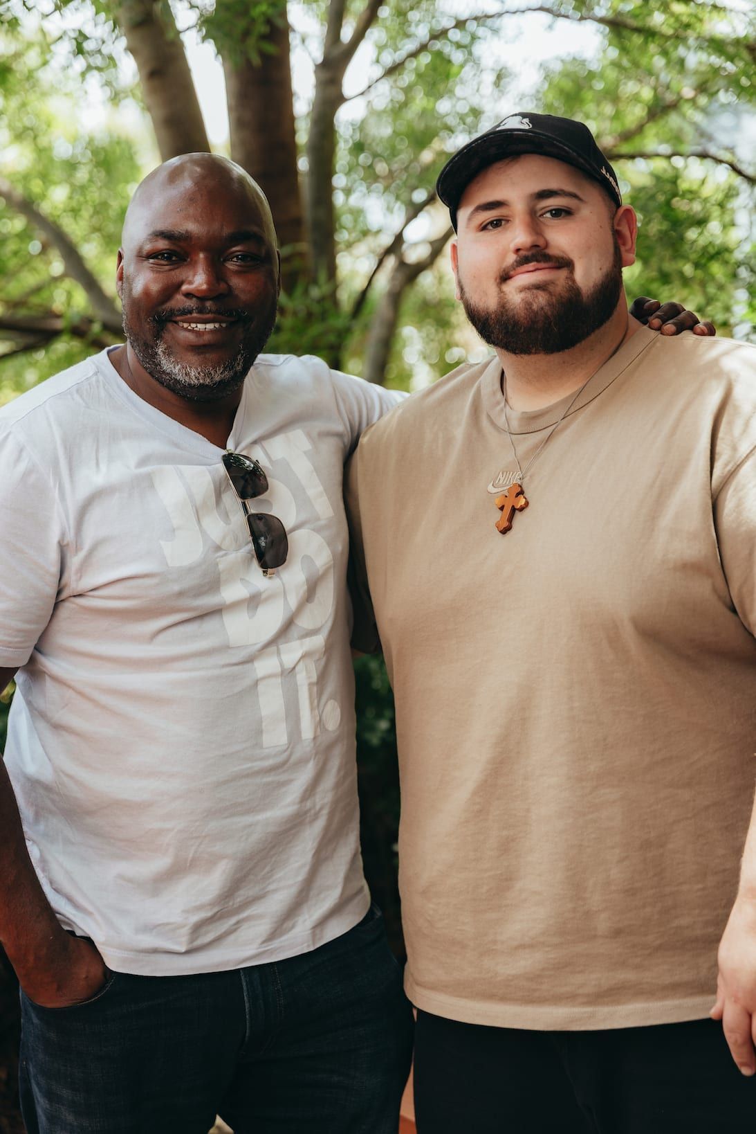 Two men pose smiling outdoors. One in a white shirt, the other in a tan shirt and black cap.