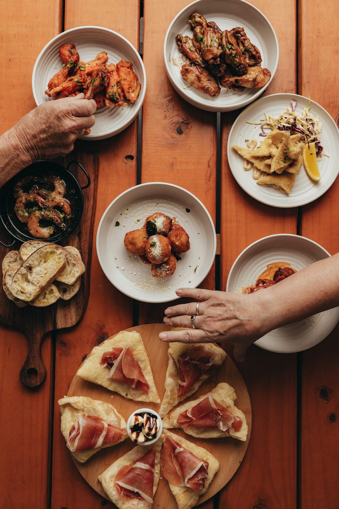 Assortment of appetizers: wings, arancini, flatbread, dips, and hands reaching for food on a wooden table.