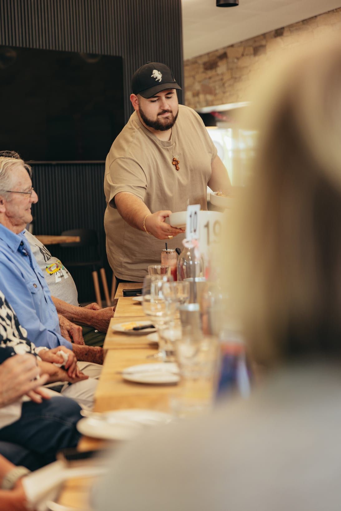 Waiter serving food to a seated group at a restaurant; light-colored clothing, wooden table.