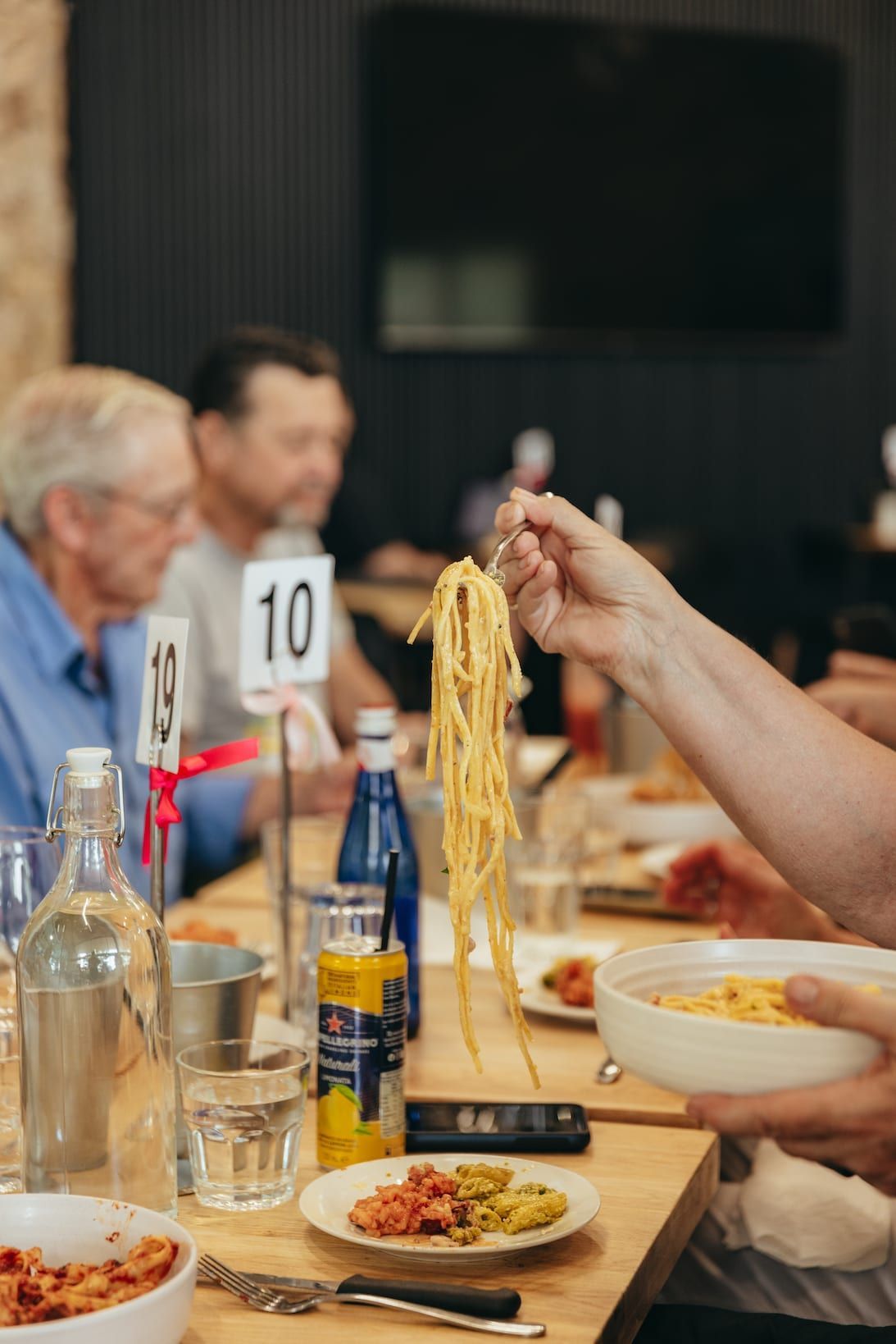 Person holding spaghetti above a table set for a meal. Other diners are visible, along with water bottles, food plates.