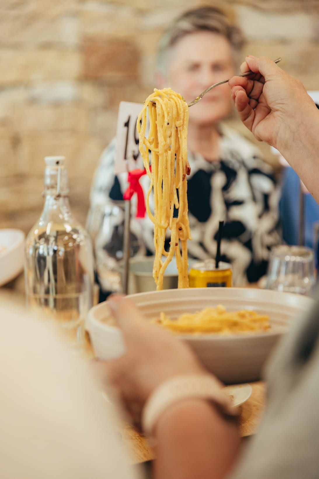 Person twirling pasta on a fork over a bowl, another person seated in background at a table.