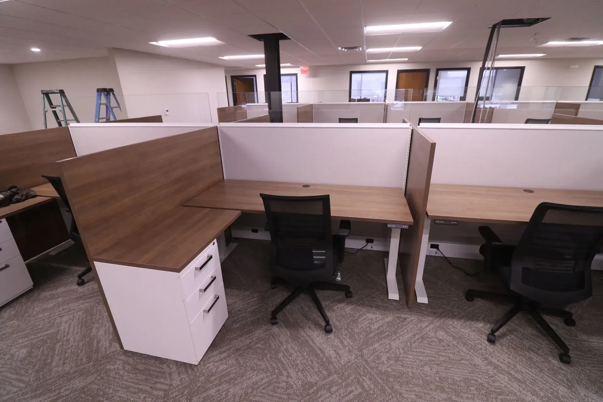 Office cubicles with desks, chairs, and dividers in a bright, empty room. The desks are wood-toned and white.