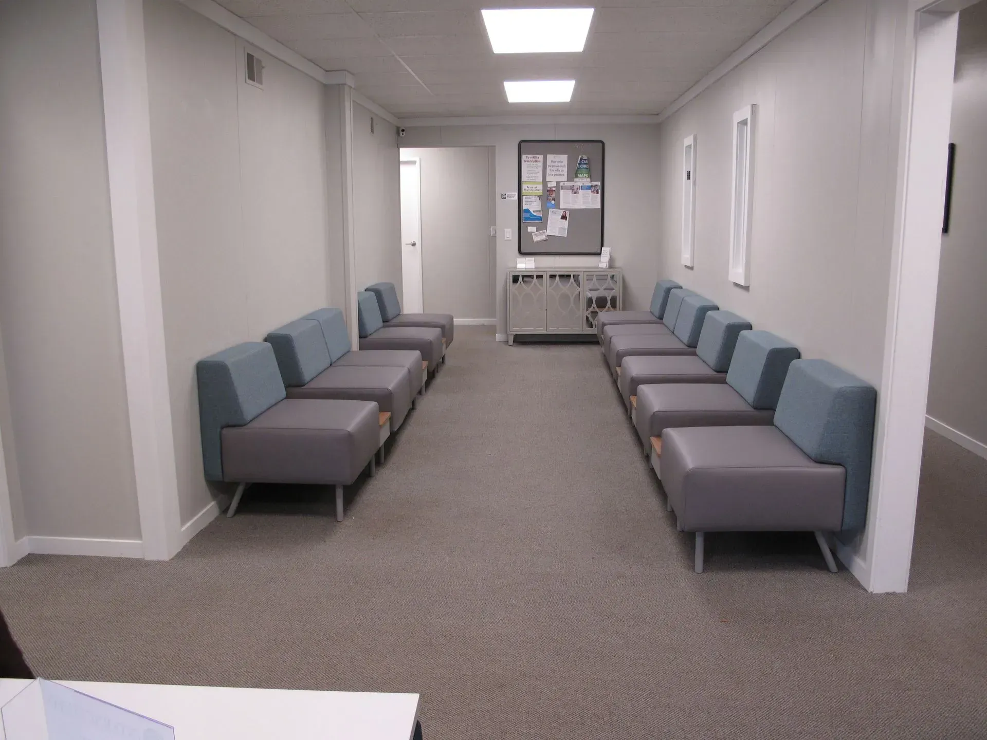 A long, narrow waiting room with grey couches lining each side. A bulletin board and door are visible at the end.