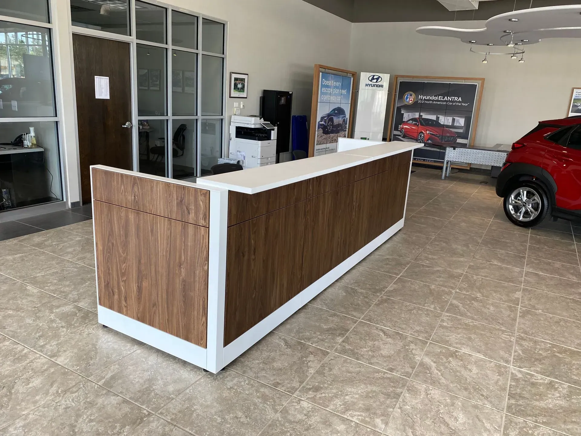 A wooden and white reception desk in a car dealership showroom. A red car is partially visible.