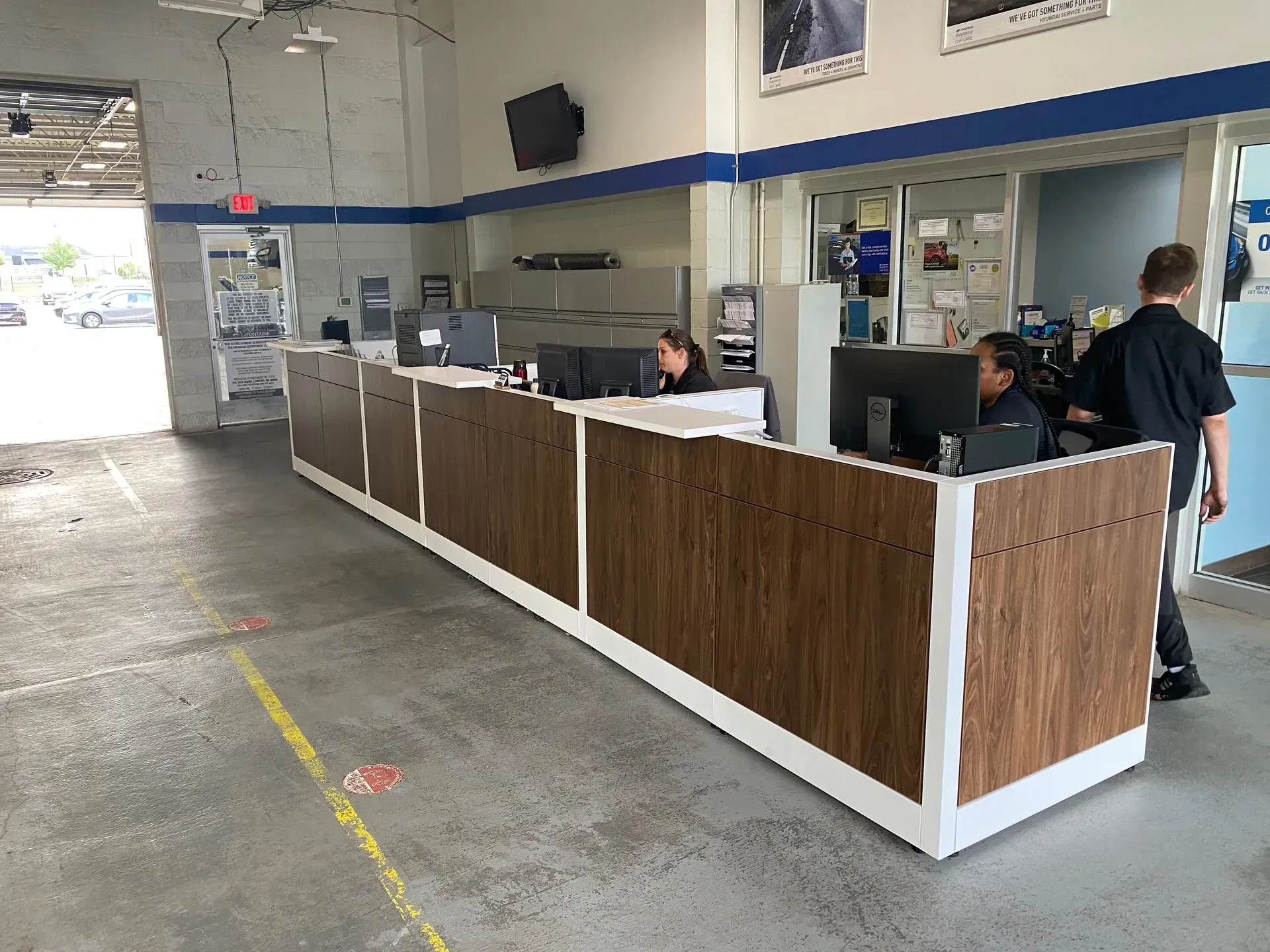 Long reception desk with people in an auto shop setting. Two workers assist customers; one with dark skin sits at a computer.