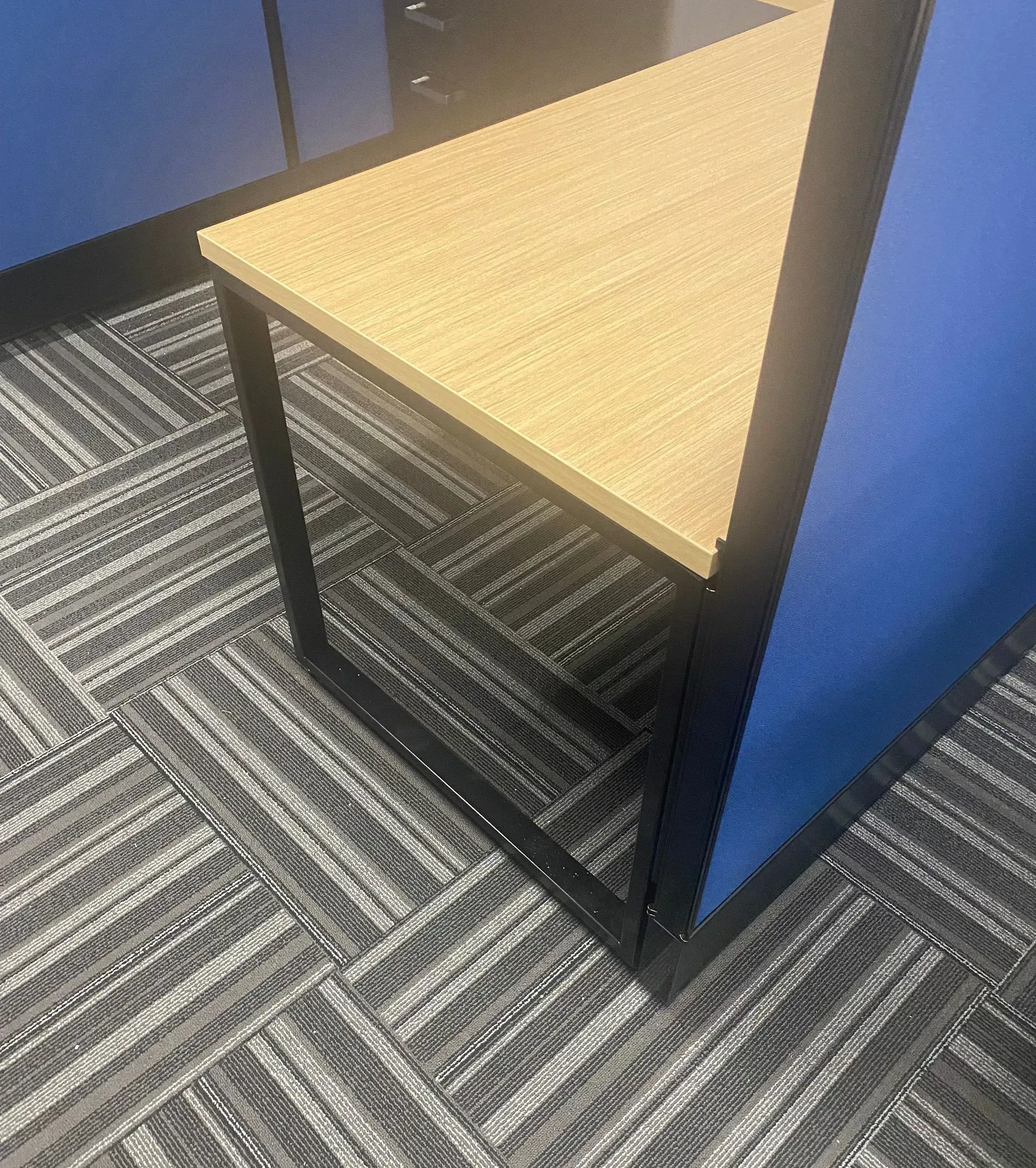 A close-up of an office cubicle corner. A wooden triangle desk rests on a black frame, with blue walls.