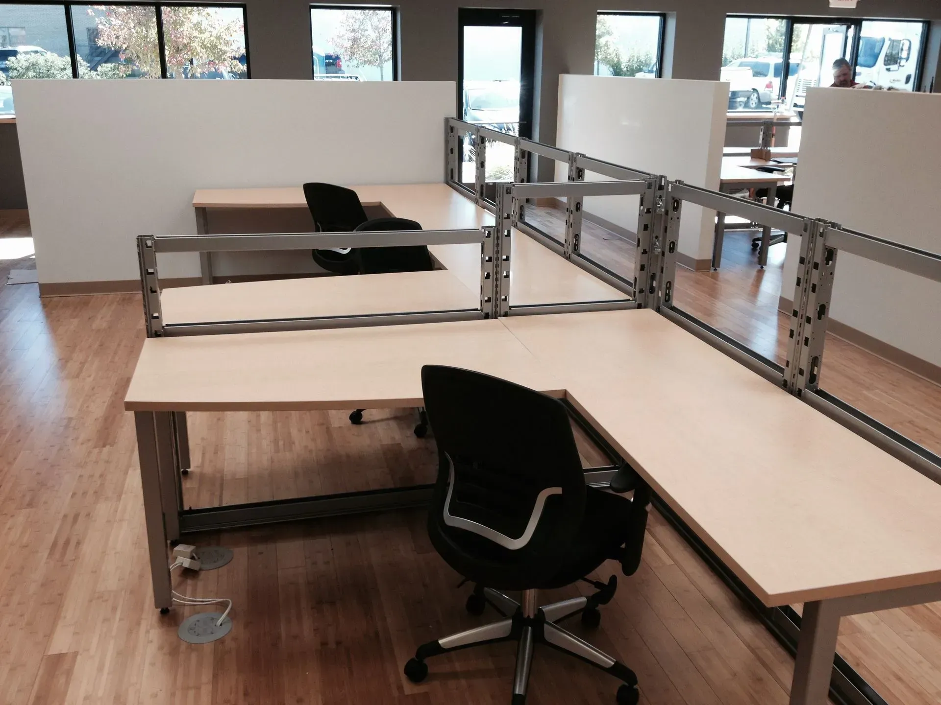 Office space with light wood desks, black chairs, and metal dividers on a wood floor. Large windows are in the background.