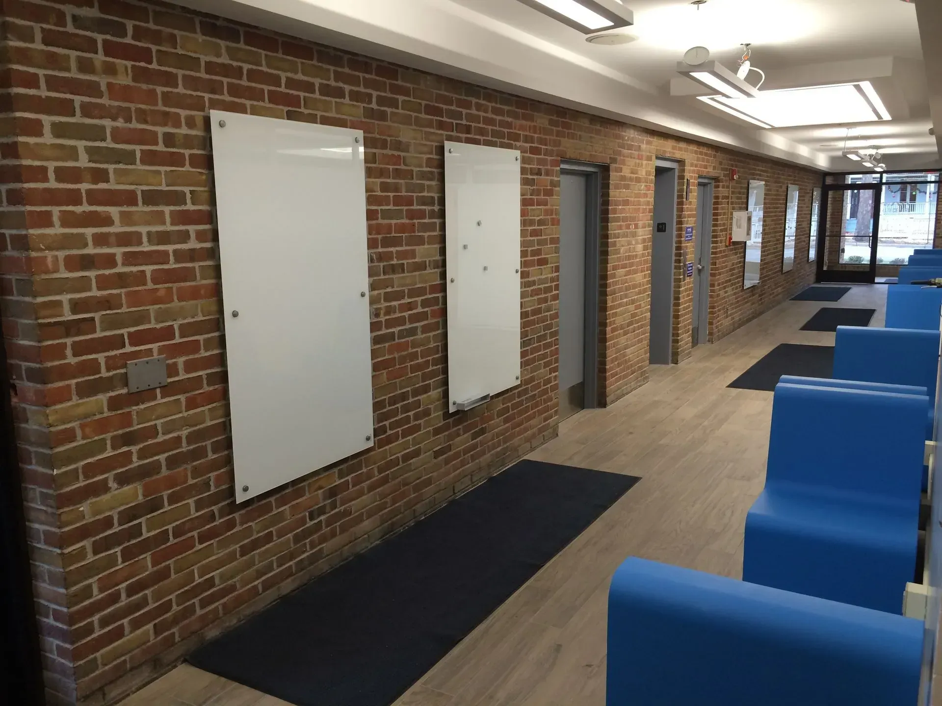 A long hallway with exposed brick walls, whiteboards, blue chairs, and gray doors. Sunlight streams in from the far end.