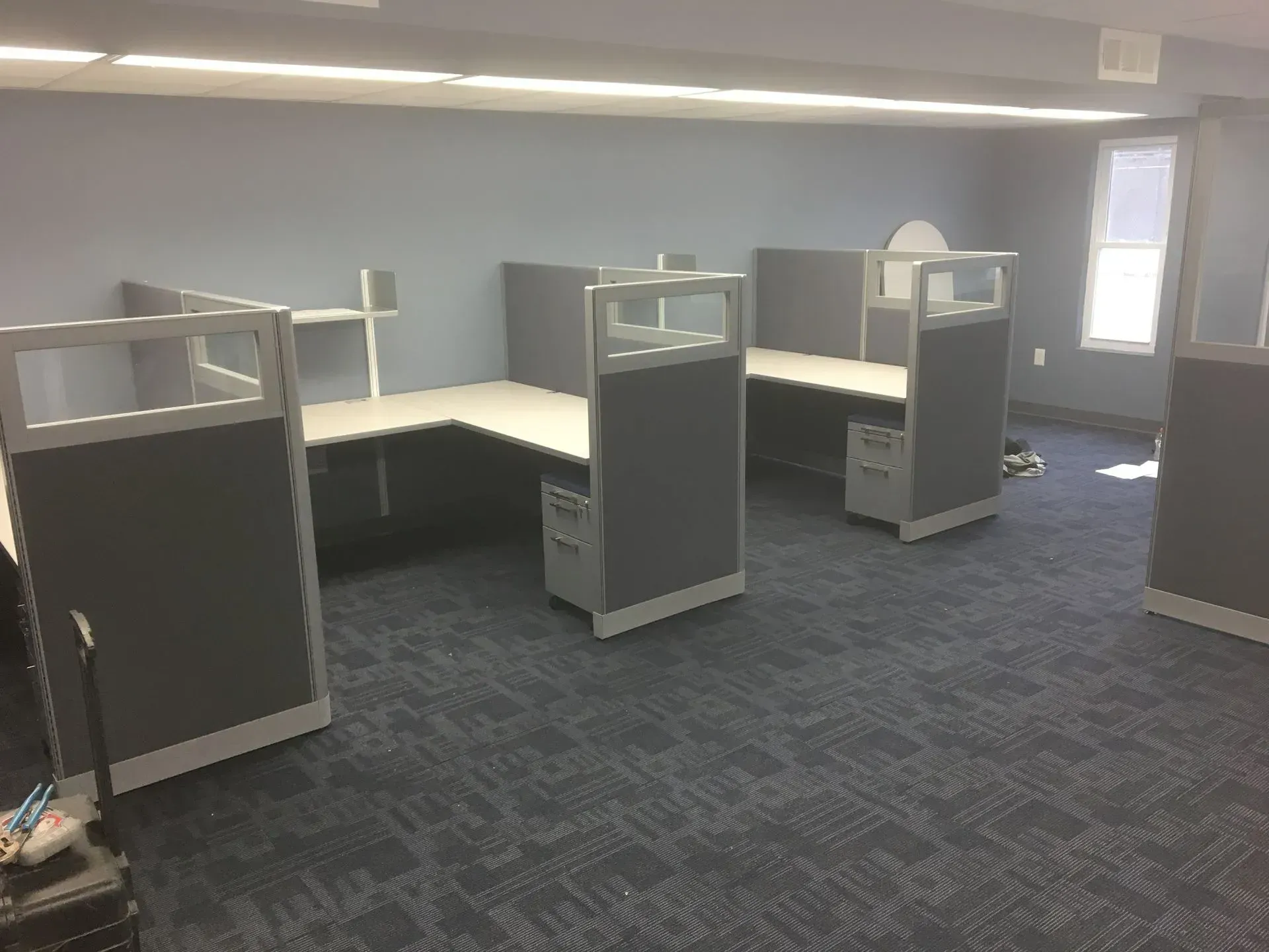 Office cubicles with gray fabric panels and light gray desks on blue patterned carpet.