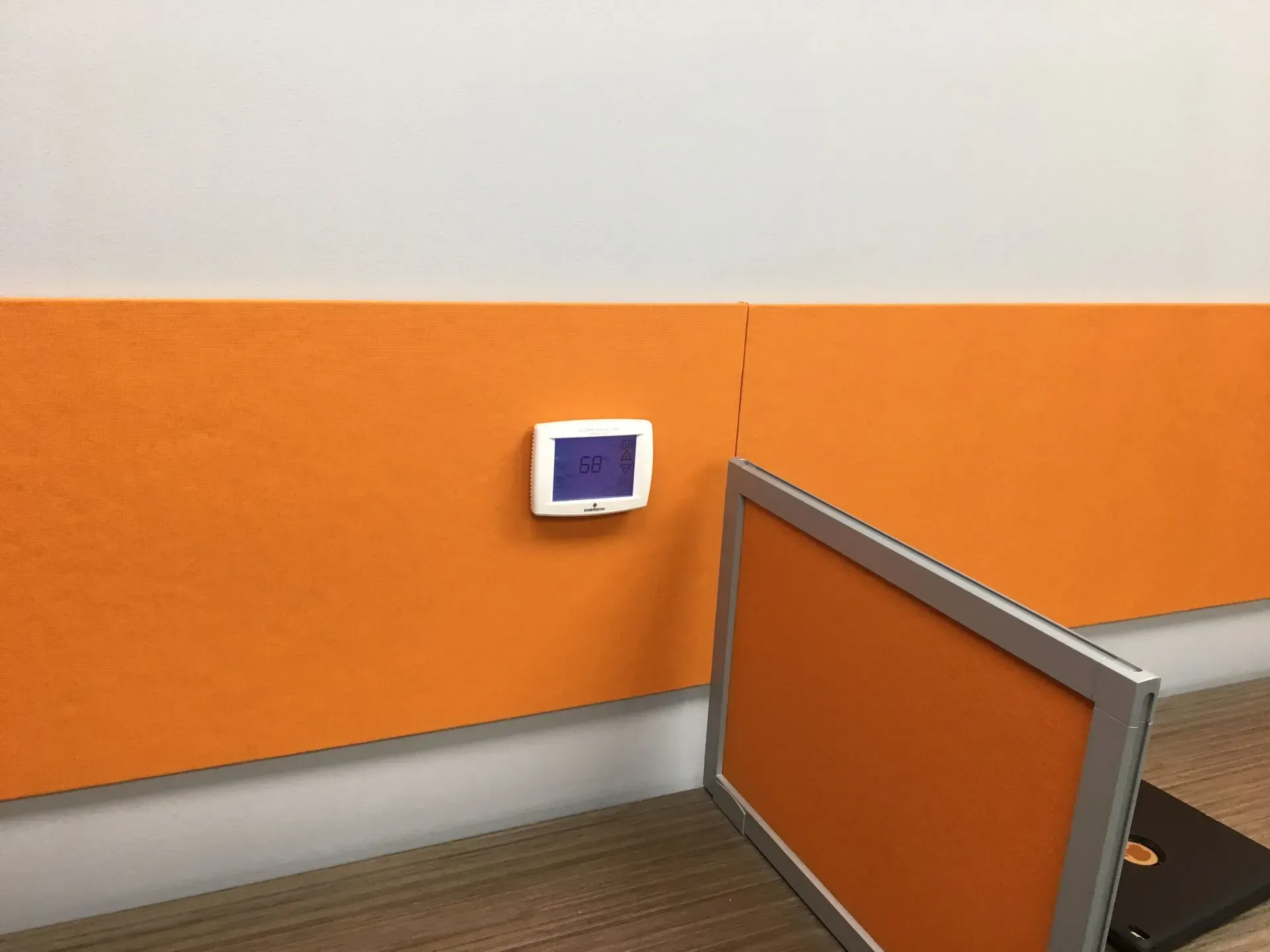 A white thermostat with a blue screen is mounted on an orange wall above a desk. A computer monitor is on the desk.