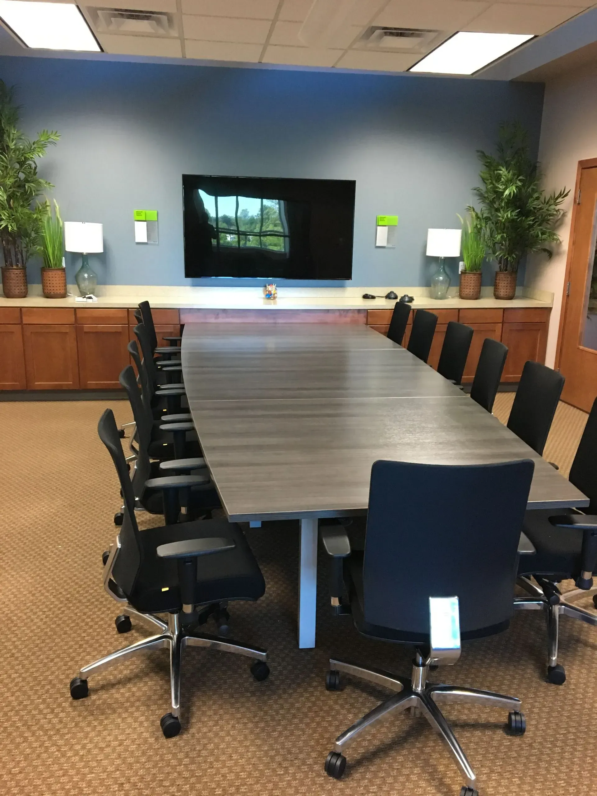 Conference room with long gray table, black chairs, and large screen on a blue wall.