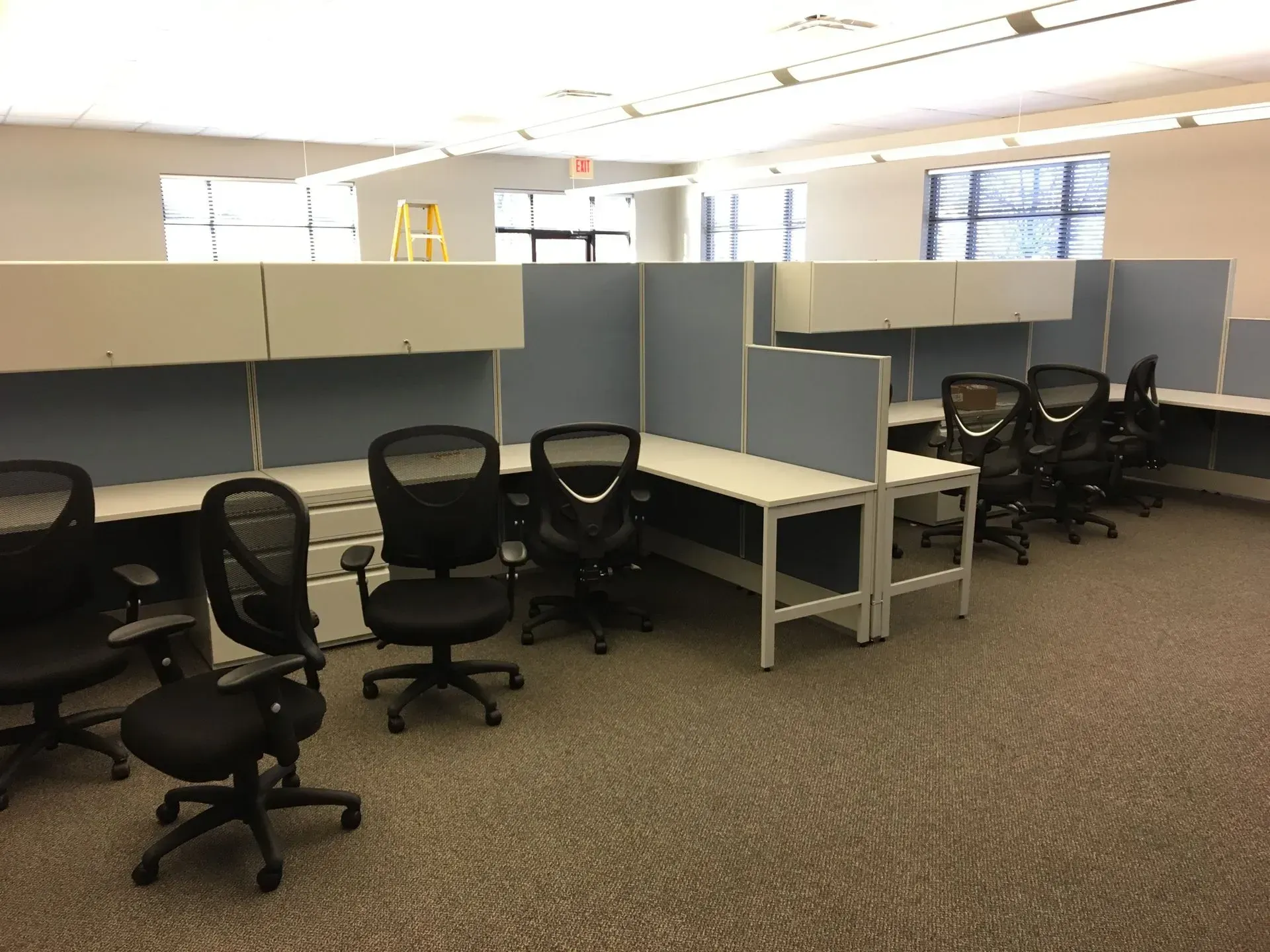 Empty office cubicles with black chairs and light gray desks and cabinets. Cubicle walls are light blue and white.