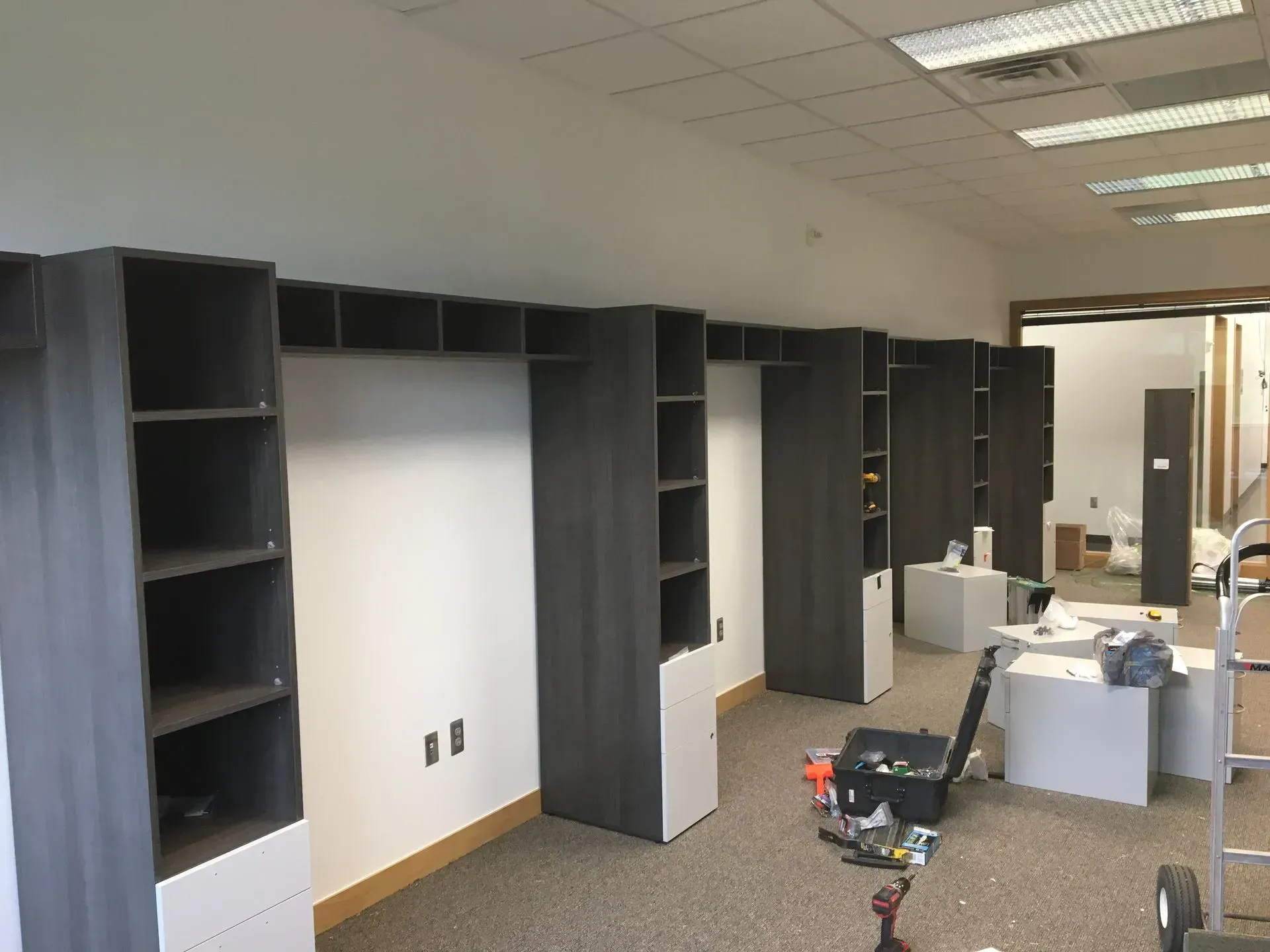 Shelving units under construction in an office space. Dark gray shelves are mounted against white walls with visible wiring.