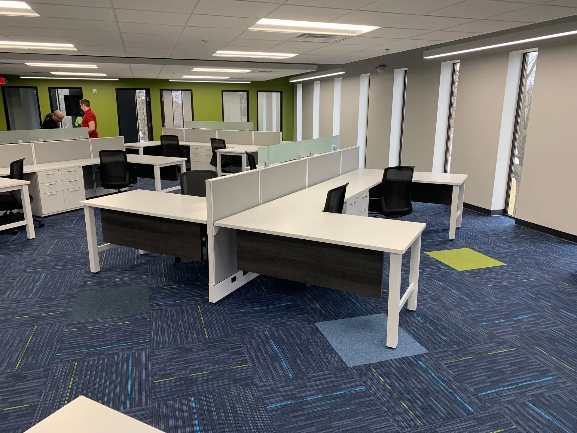 An office space with several cubicle desks and colorful carpet. A couple of people stand in the background near a green wall.