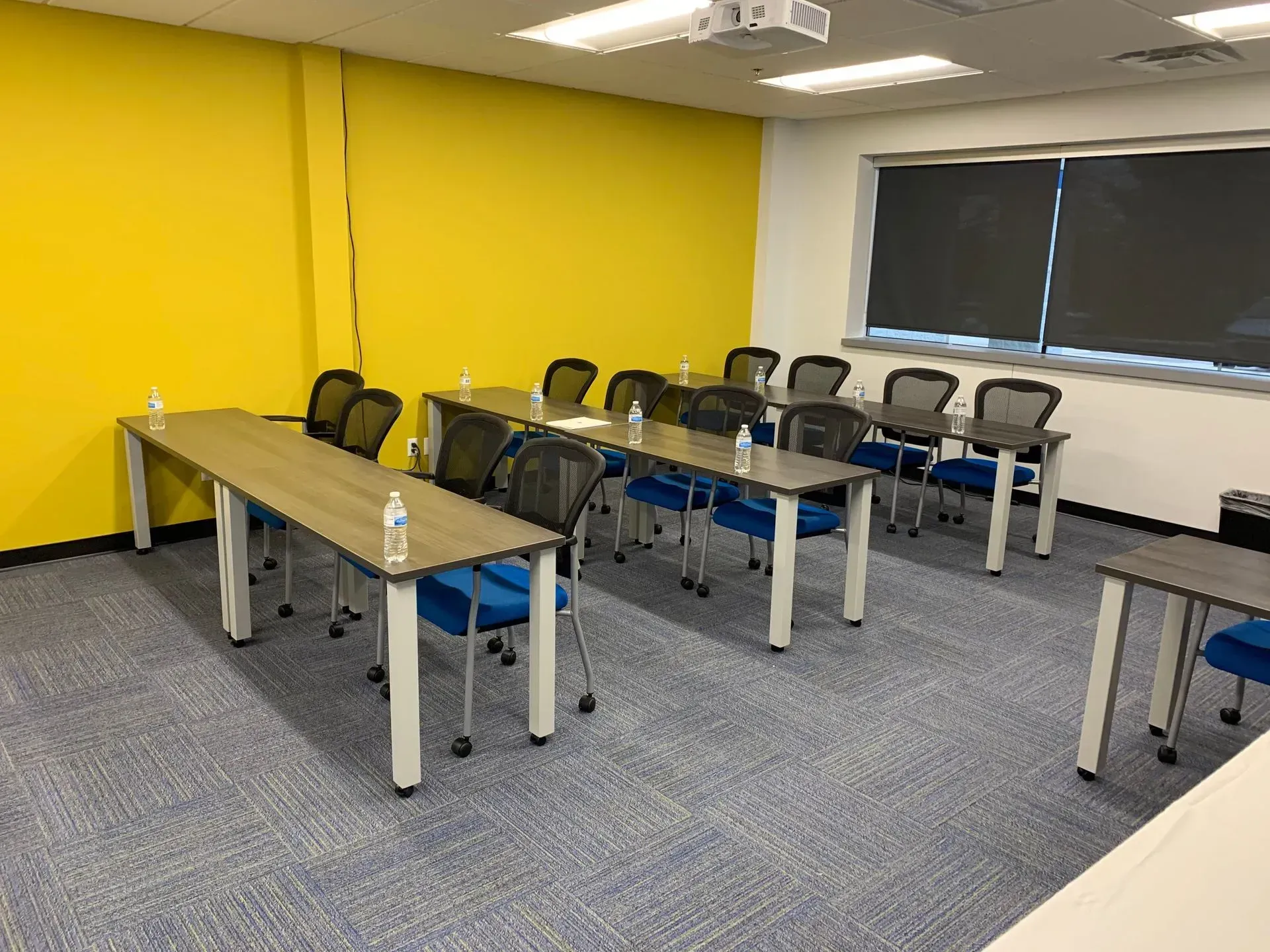 Classroom with long tables, blue chairs, and a yellow wall. There are water bottles on the tables, and a window with a blind.