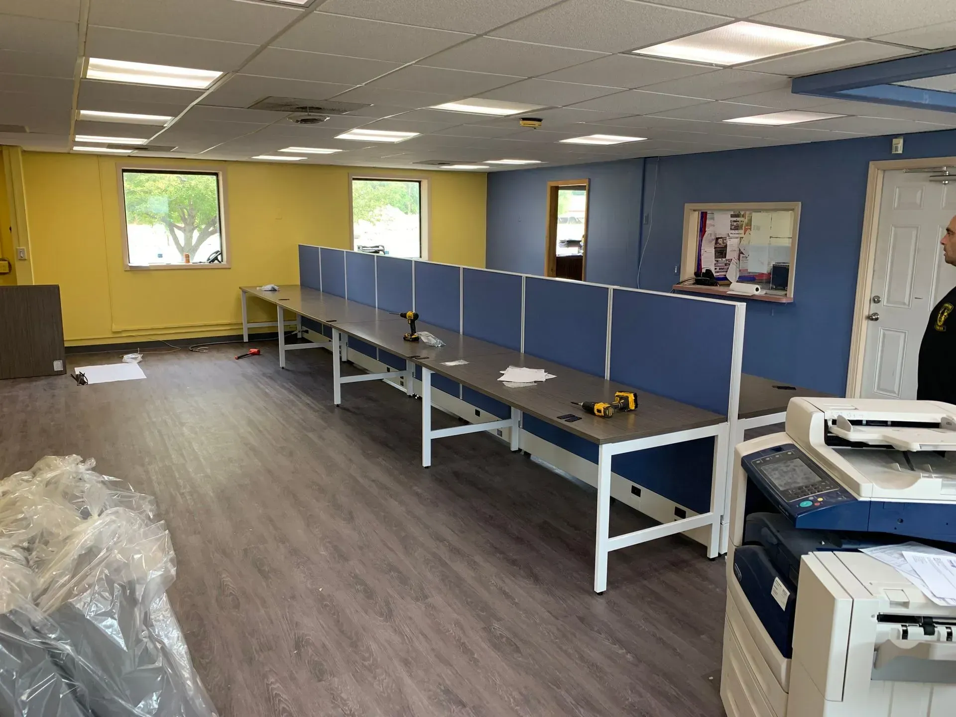 An empty office space with long desks, blue dividers, and a mix of yellow and blue walls. A person stands near a printer.