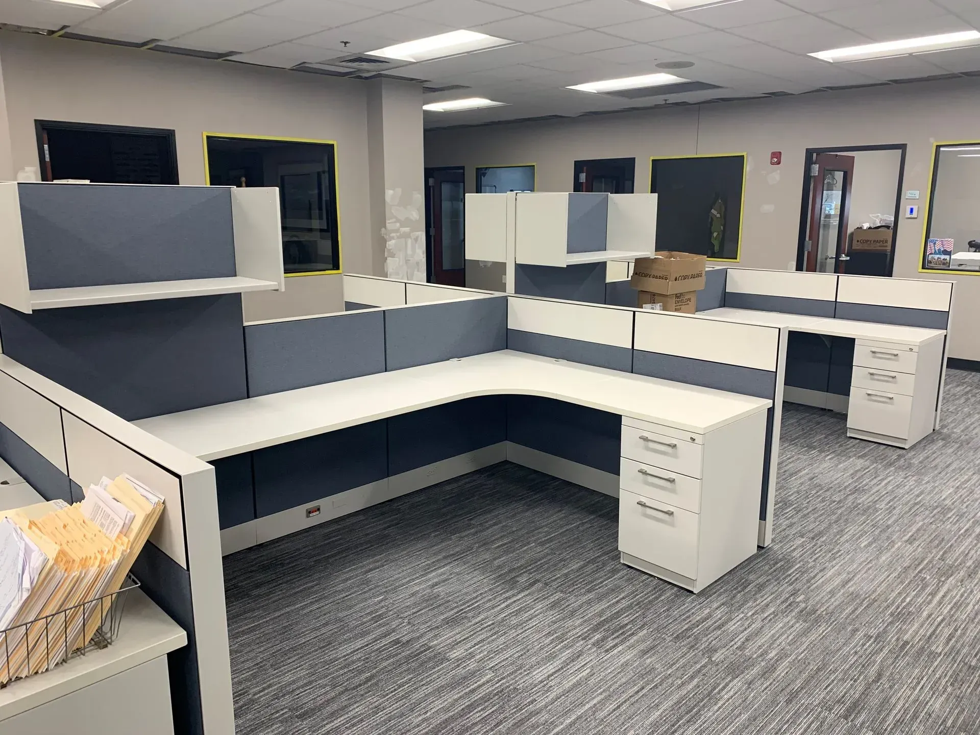 An office space with several gray and white cubicles. Some cubicles have overhead storage. The floor is gray carpet.
