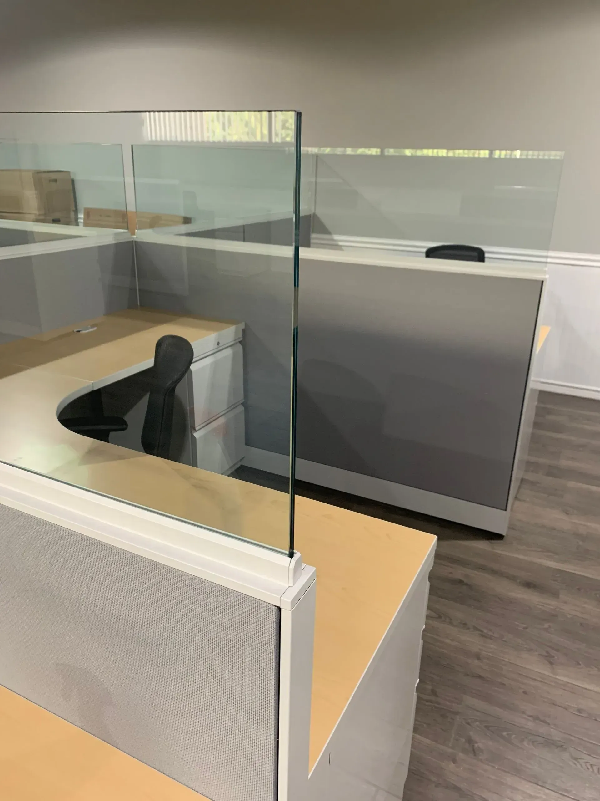 Cubicle with glass partition. Light-colored office space with gray cubicle walls, wooden desks, and a black chair visible.