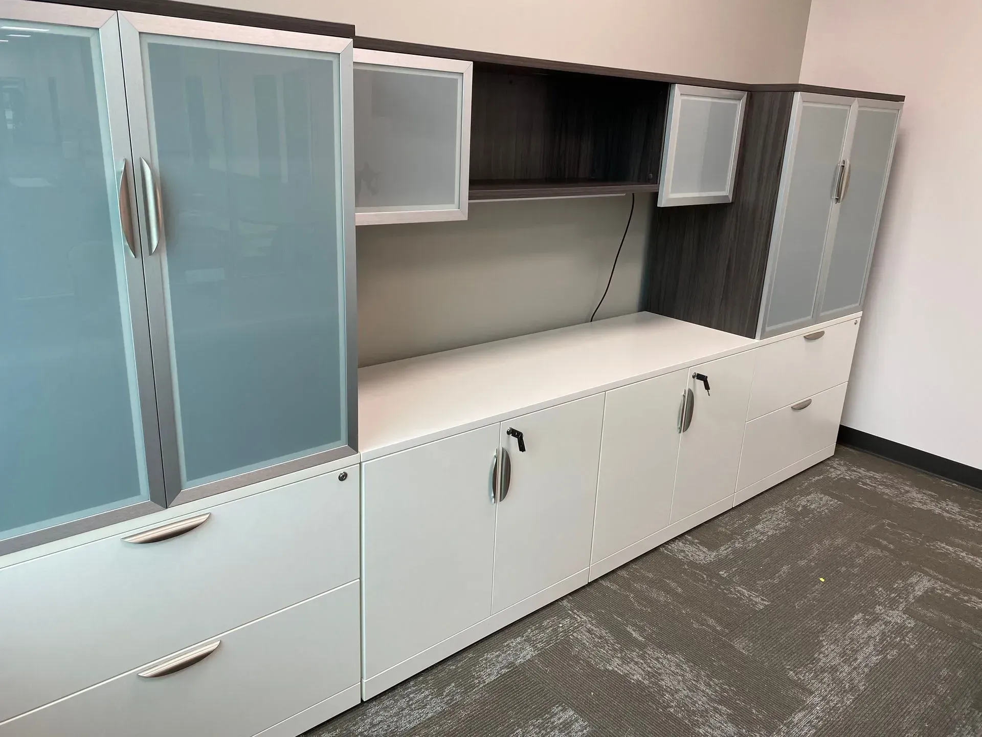 Office storage unit with gray and white cabinets, glass doors, and a black shelf above a white desk area.