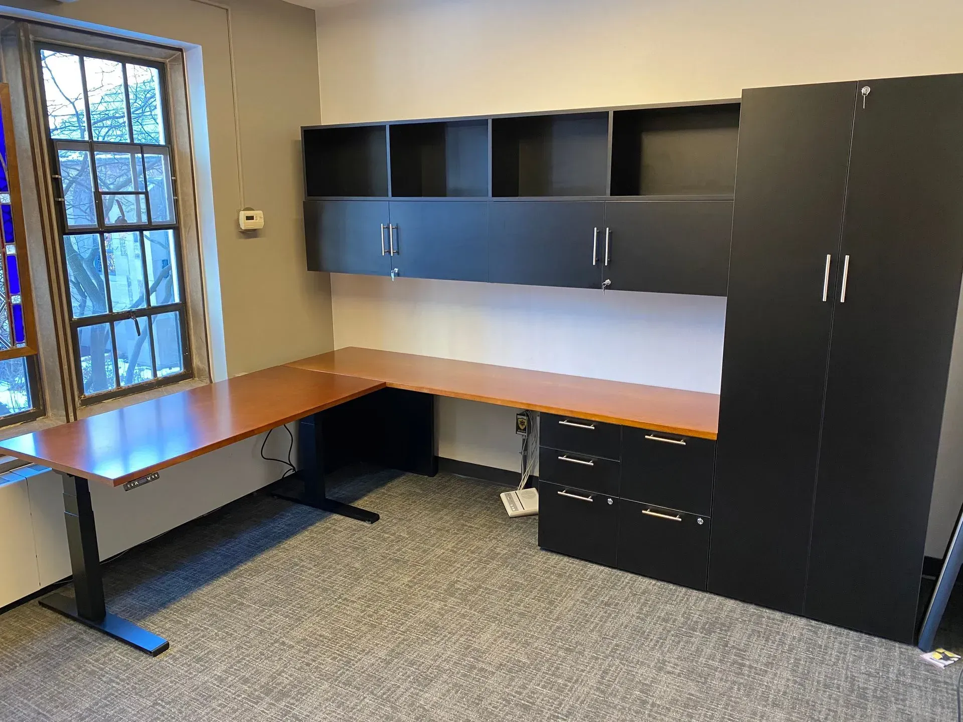 Office setup with a wooden L-shaped desk, black cabinets, shelves, and a large window.