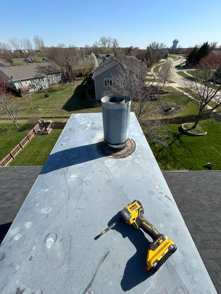 Rooftop view with a chimney and a yellow power drill; houses and green lawns in the distance under a clear blue sky.