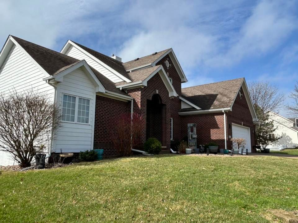 Two-story brick and white-sided house with brown roof on a grassy lawn under a partly cloudy sky.