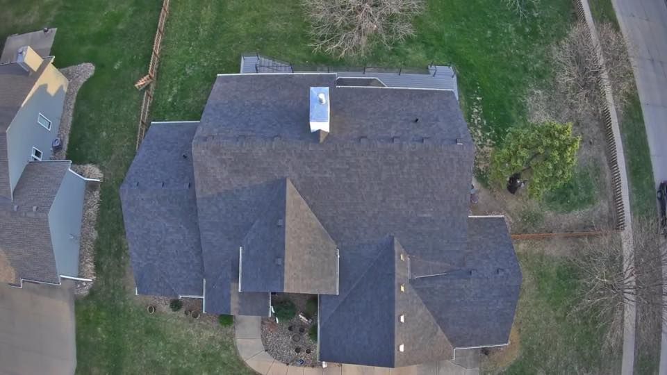 Overhead view of a dark shingled roof with a chimney, surrounded by grass and other houses.