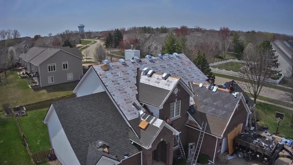 Aerial view of a house roof being replaced; shingles partially removed, ladders visible, surrounded by green grass and other homes.