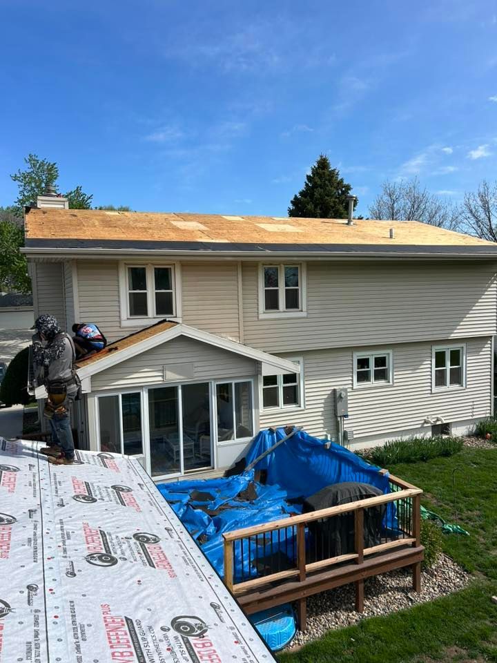 Roofers working on a house; blue tarp on deck, wood siding, sunny day.