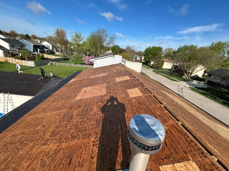 Roof with exposed plywood, shadow of photographer, crew working on roof. Suburban setting, sunny day.