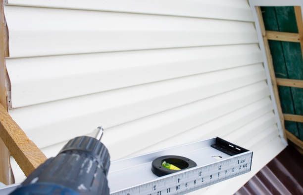 A person uses a drill to attach white siding to a building, with a level for guidance.