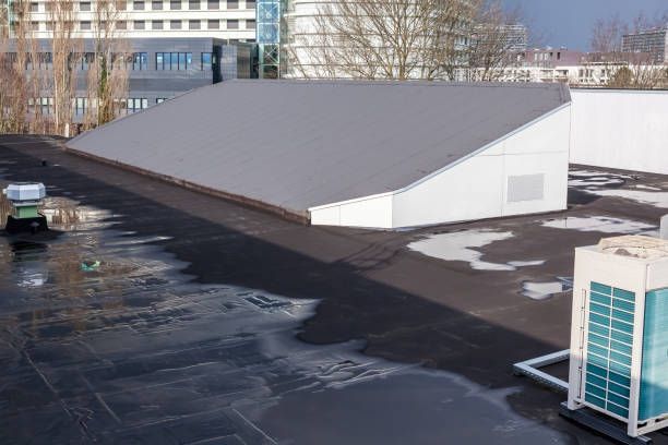 Flat black rooftop with water puddles; sloped gray roof structure on white enclosure; distant buildings.