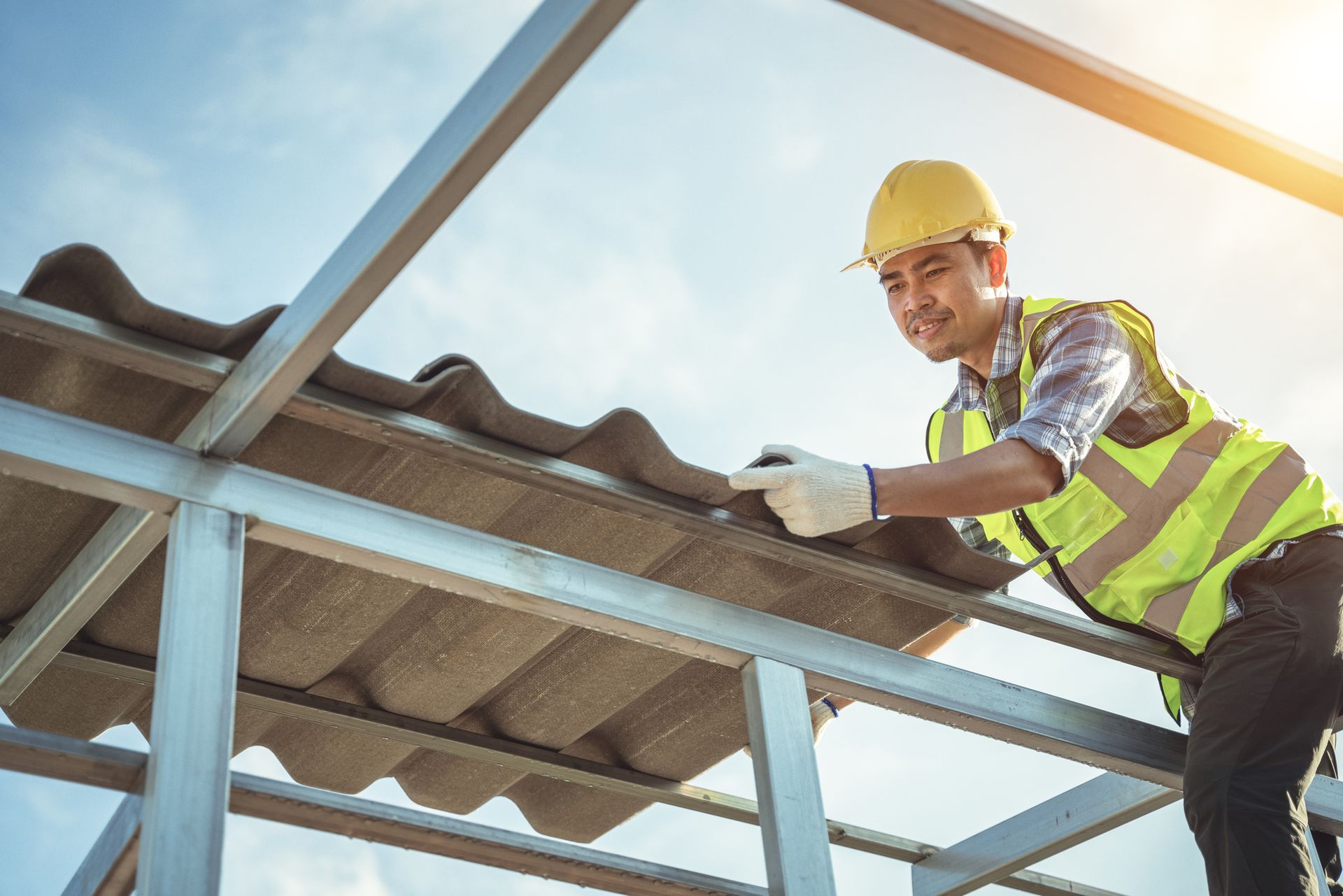 Roofer in a yellow helmet, placing roof tiles on a metal frame, wearing a yellow safety vest, blue sky.