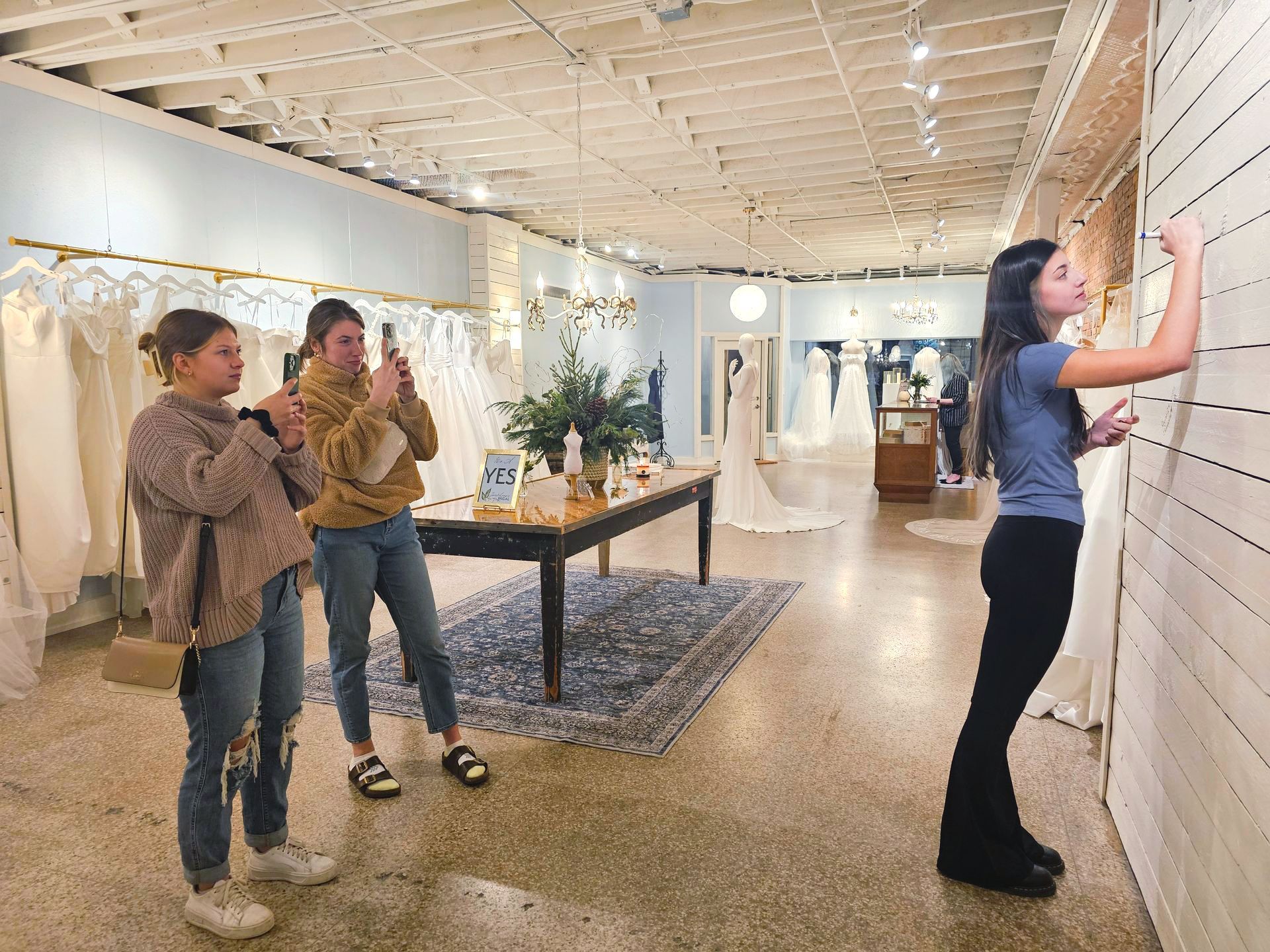 Three young women in a bridal shop: two taking photos of a bride as she points at a wall. The shop has dresses on display.