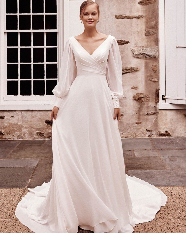 A woman in a white wedding dress with long sleeves is standing in front of a building.