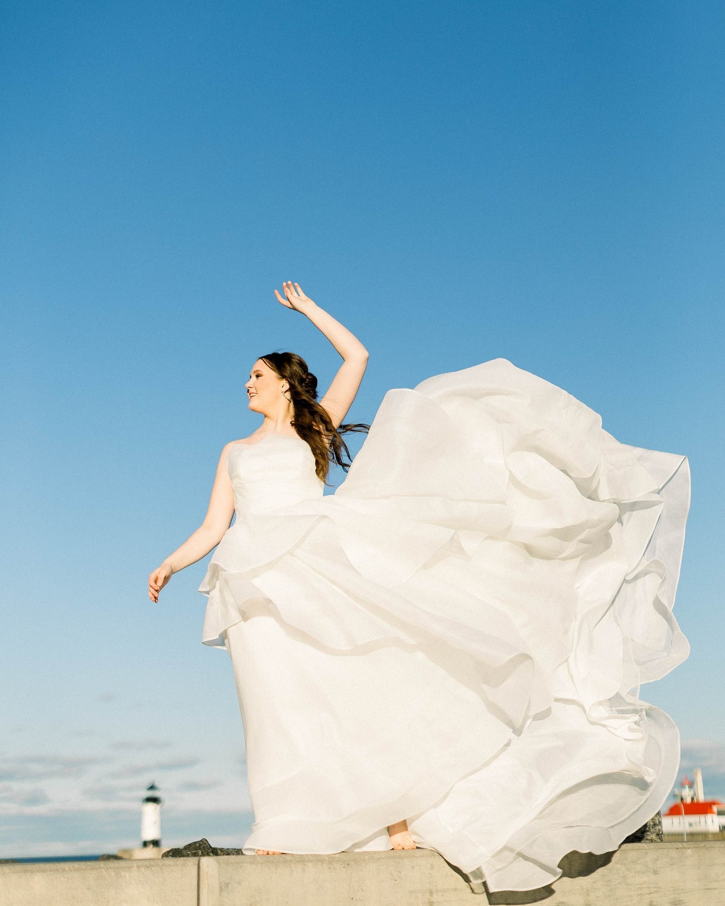 Bride in a flowing white gown dances on a stone wall under a bright blue sky, with a lighthouse visible in the background.