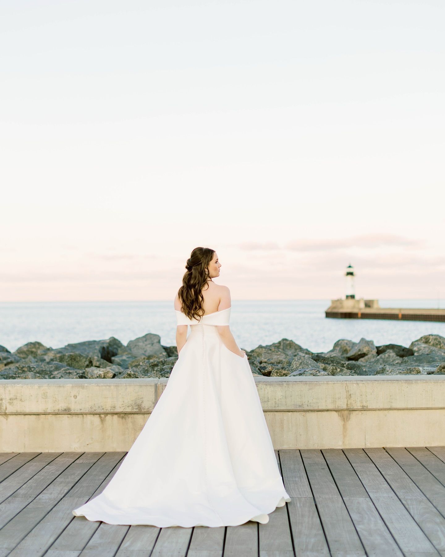 Bride in a white gown stands on a wooden deck overlooking a calm ocean with a lighthouse in the distance.