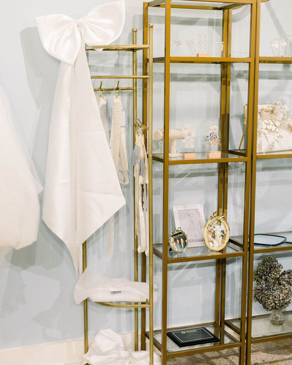 A gold display rack in a boutique showcases a large white bow, hanging garments, and jewelry on glass shelves against a pale blue wall.