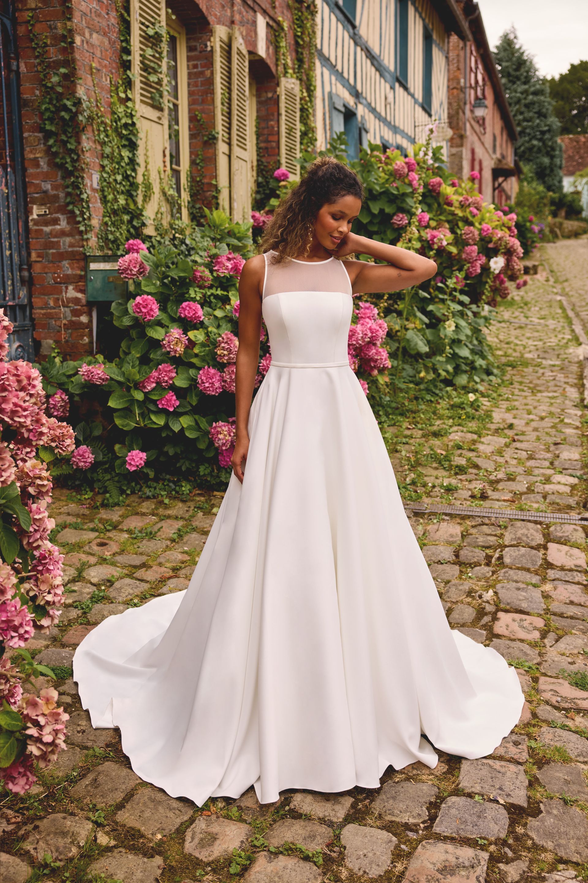 Woman in a white wedding dress stands outside. She has curly hair, smiles, and the setting includes flowers and a stone building.