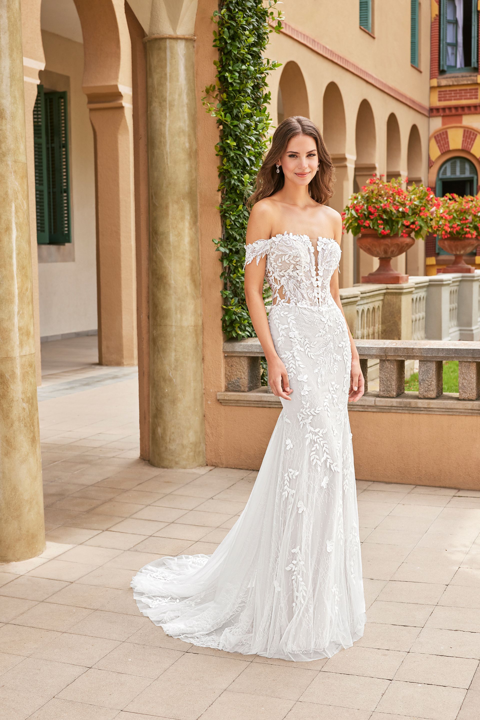 Woman in a white off-the-shoulder wedding dress stands outdoors in front of a building with columns and arches.