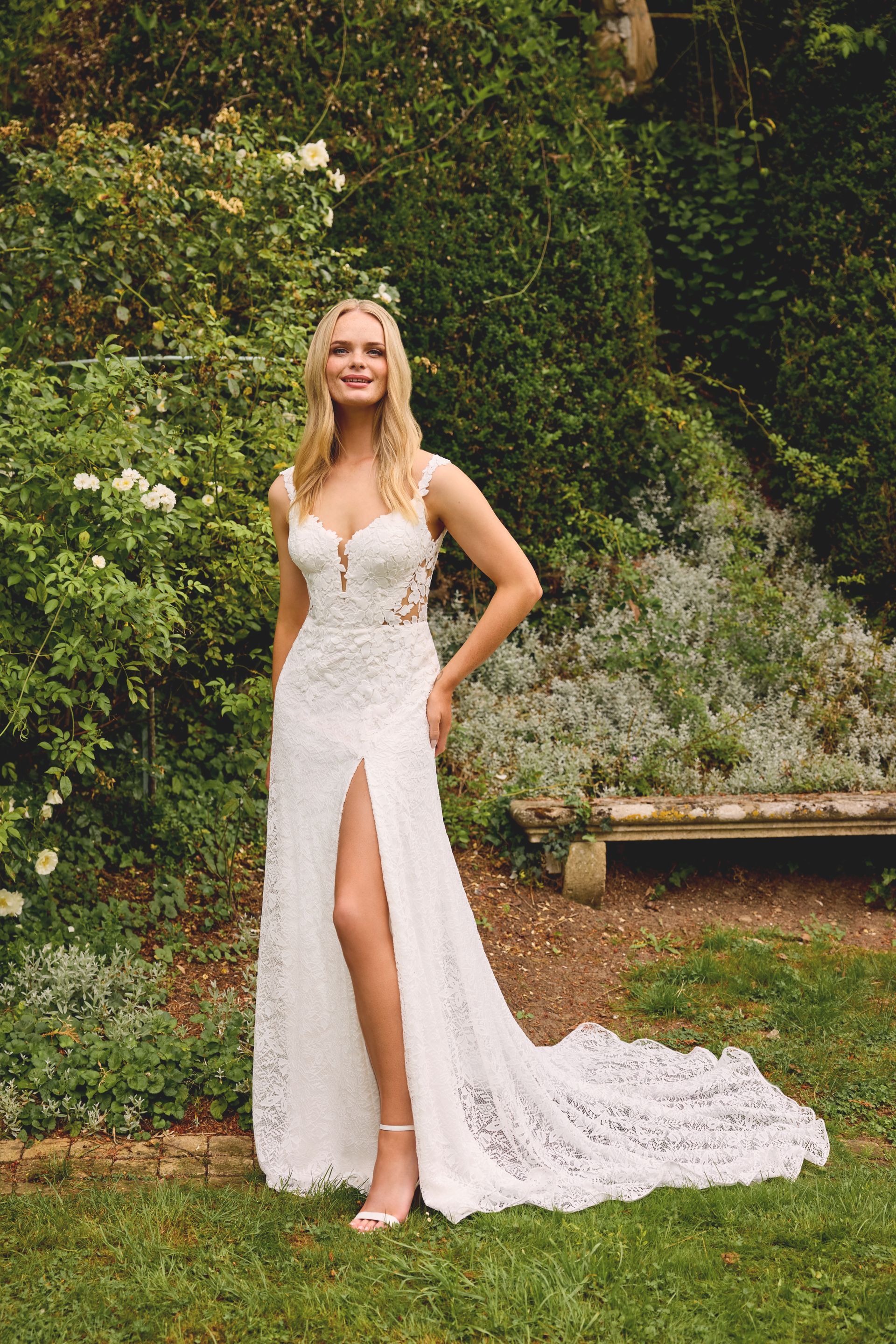 Blonde woman in off-the-shoulder wedding gown stands on cobblestone street, surrounded by flowers and historic buildings.